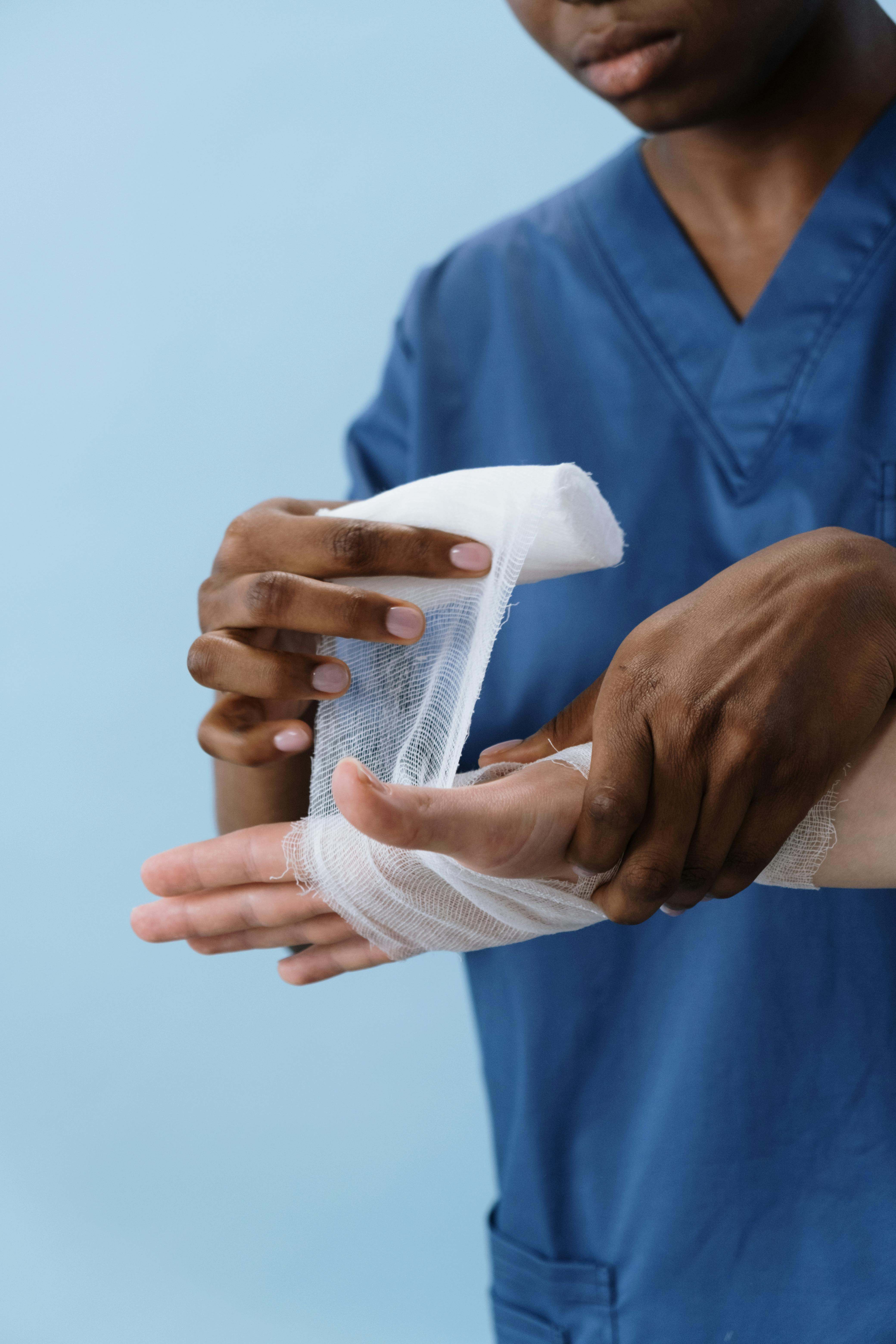 A clinician in blue scrubs carefully wrapping a gauze bandage around a patient’s hand.