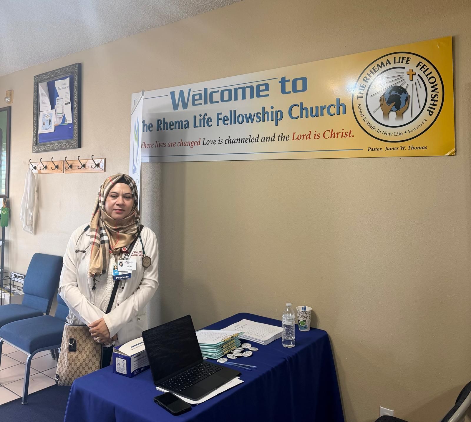 Dr. Rizvi at the health fair welcome table beneath The Rhema Life Fellowship Church sign